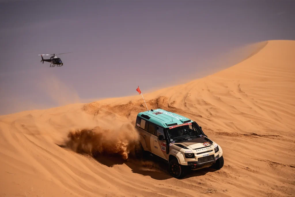 defender dakar cresting a dune followed by a helicopter