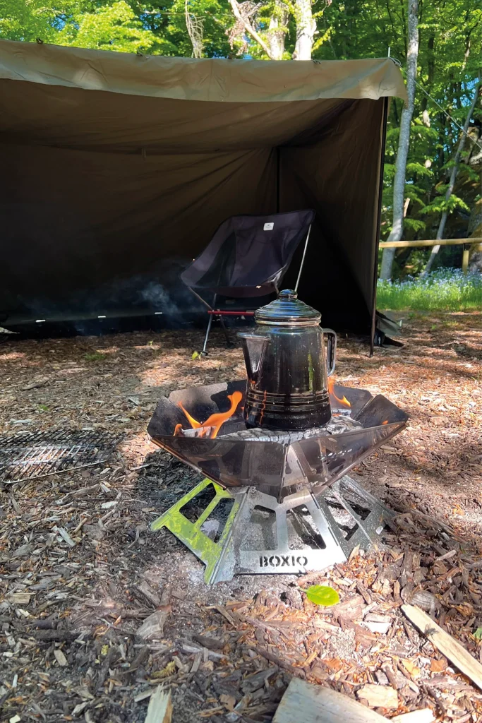 Enamel coffee pot heating over a BOXIO FIRE fire pit beside a campsite tarp during an overland camping trip.
