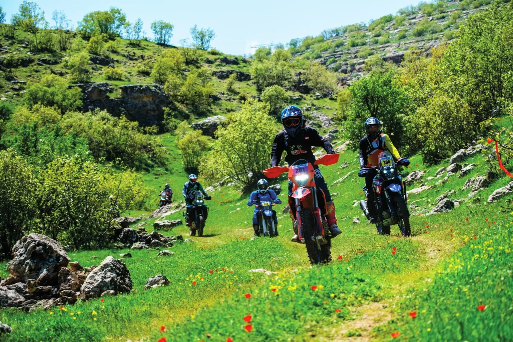 Motorcycle riders competing in the Sky Adventure Race across a green, rocky mountain trail near Savur in southeastern Turkey.