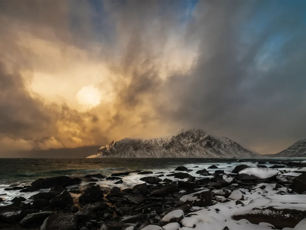 Snow-covered rocky shoreline beneath storm clouds and low winter sunlight in the Lofoten Islands.