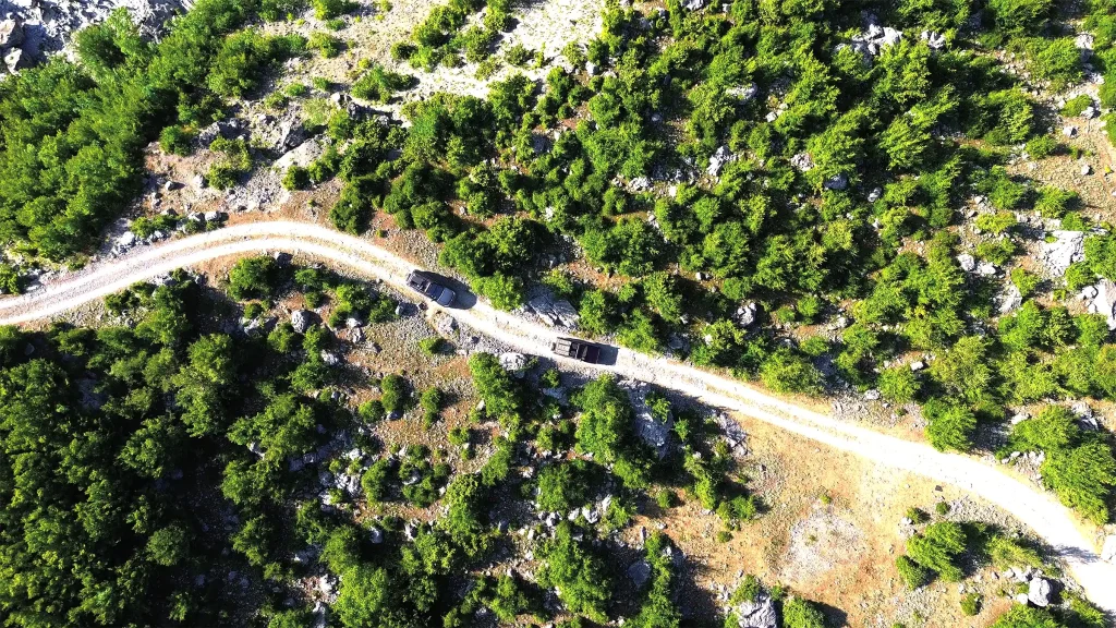 Aerial view of two overland vehicles travelling along a winding gravel road through dense green forest in Albania.