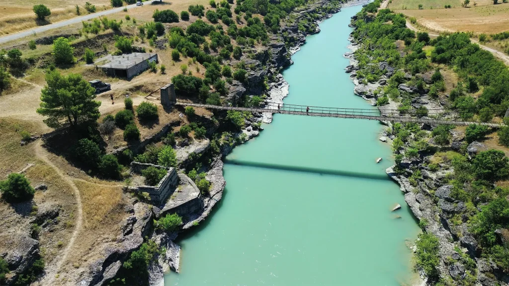 Aerial view of a suspension bridge crossing a striking turquoise river in the Albanian countryside.