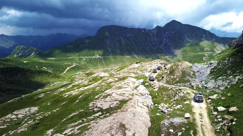 Convoy of off-road vehicles crossing a remote mountain track beneath dramatic storm clouds in Albania.