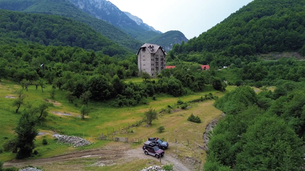 Two overland vehicles beneath a remote stone building surrounded by forested mountains in Albania.