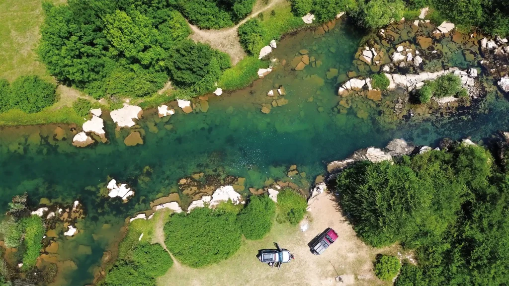 Aerial view of two overland vehicles beside a clear green river flowing through forest and rocks in Albania.