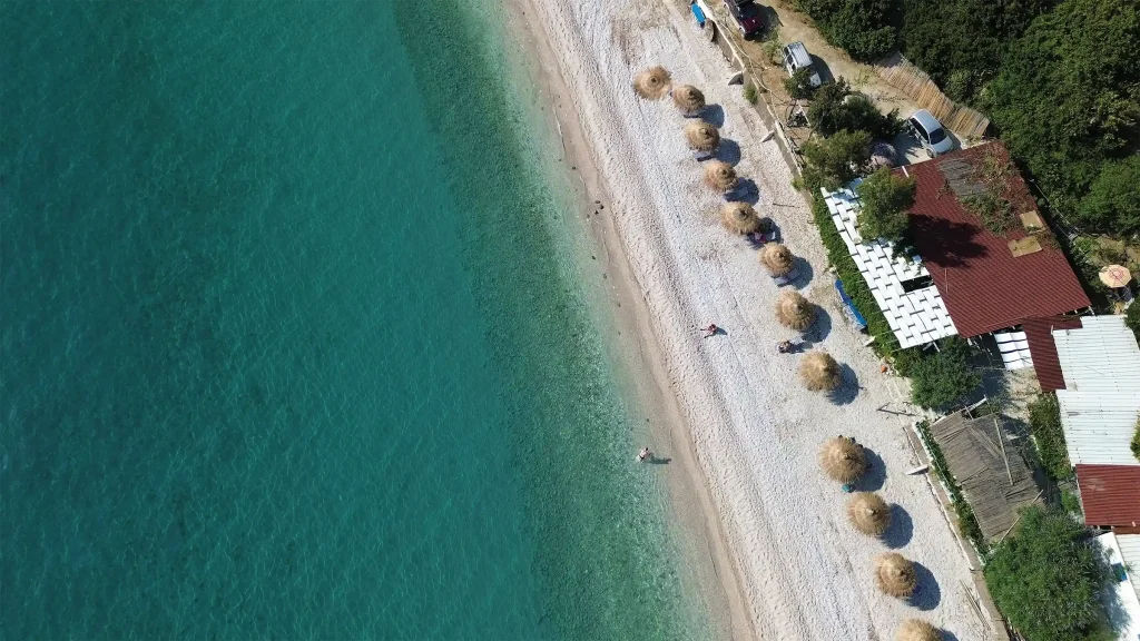 Aerial view of a turquoise Albanian beach with straw parasols and buildings along the shoreline.