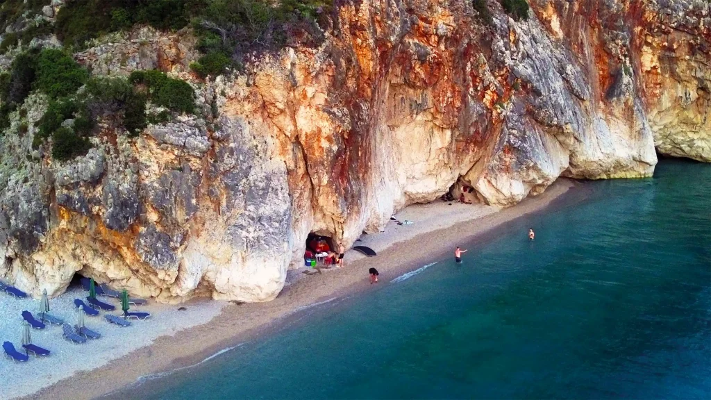 Secluded Albanian beach beneath orange and white cliffs with swimmers in the clear blue sea.