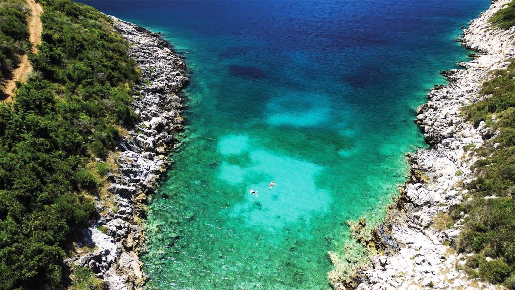 Aerial view of two swimmers in a turquoise coastal cove surrounded by rocky cliffs and dense vegetation in Albania.