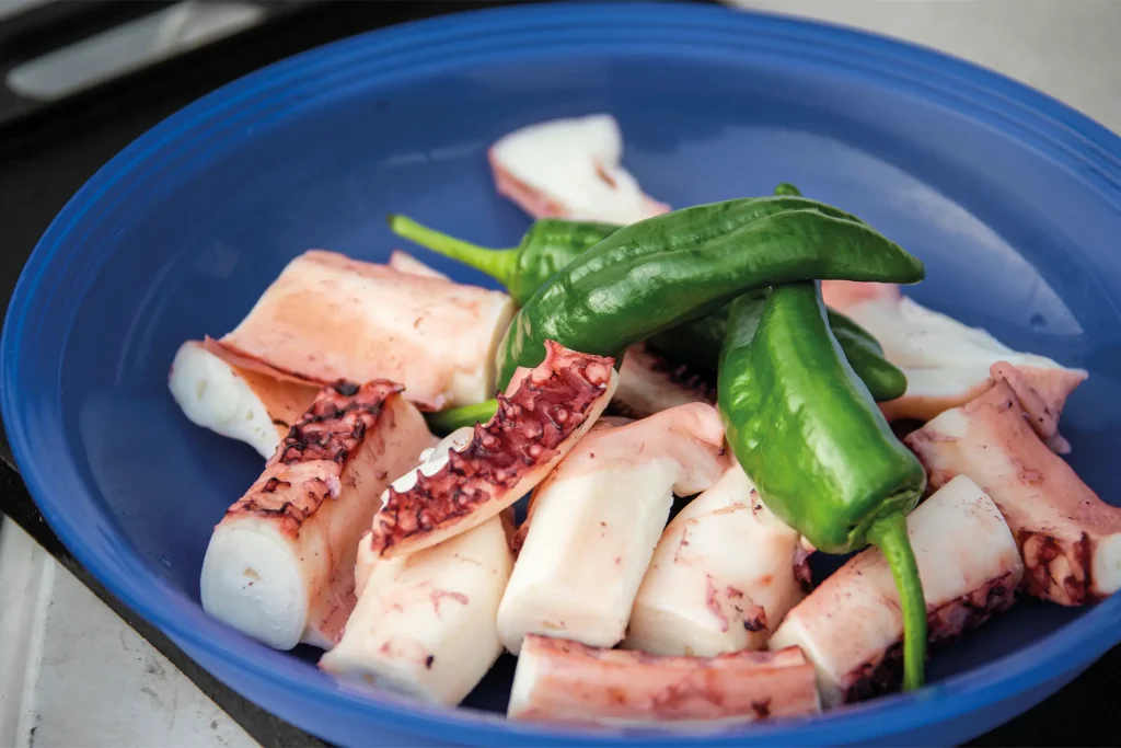 Raw octopus tentacles with green Padrón peppers in a blue bowl.