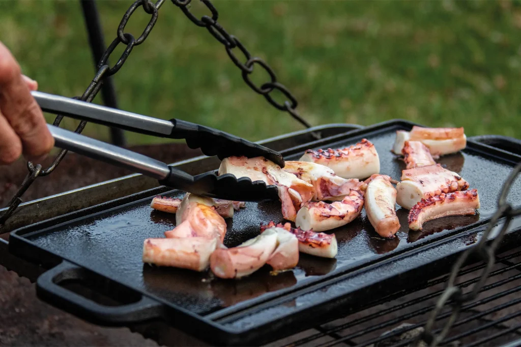 Octopus pieces being turned with tongs on a hot cast-iron plancha.