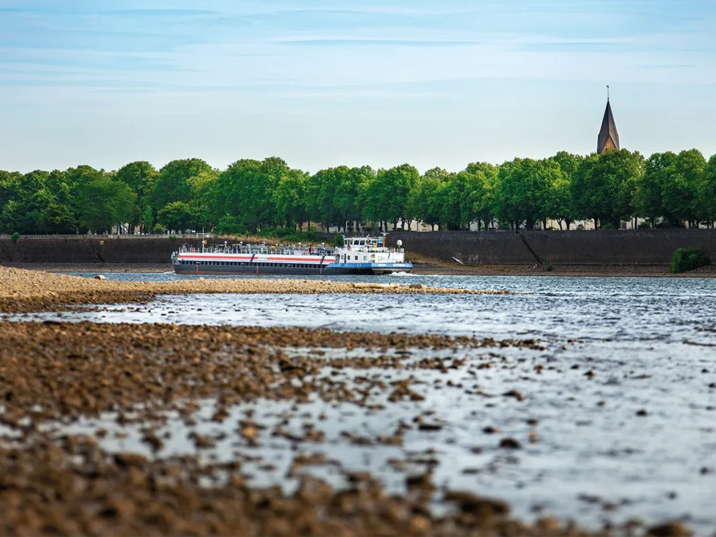 A cargo vessel travelling along the Rhine during low water, with exposed gravel banks in the foreground.