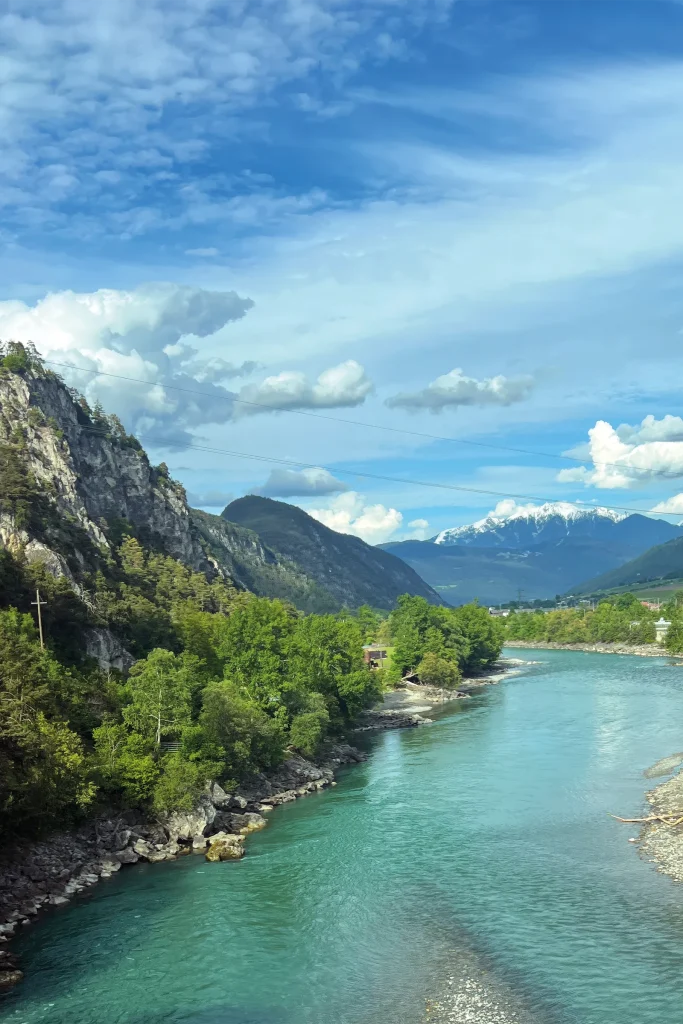 A turquoise river flowing through a South Tyrolean valley beneath wooded slopes and snow-capped mountains.