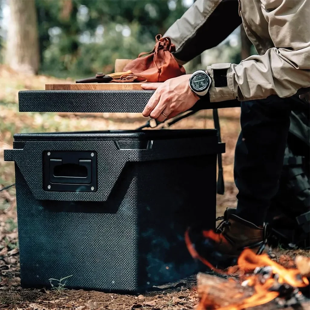QOOL passive cooler used as a camp table beside a campfire during outdoor meal preparation.