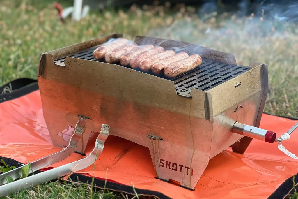 Sausages cooking on a well-used SKOTTI Gas Grill with smoke rising from the grate.